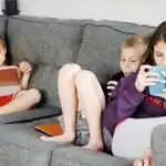 Social Media Addicts, Positive barefoot children in casual wear resting together on cozy couch and browsing tablets and smartphones